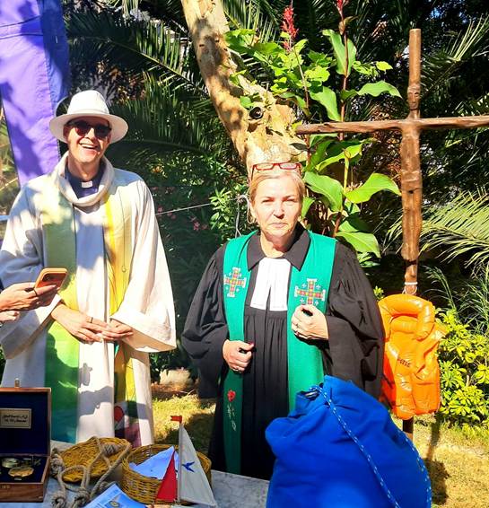 The image depicts two people standing outdoors, one dressed in a green robe and the other in a white dress, both holding books and surrounded by nature, with a cross and a blue bag on the ground.

AI-generated content may be incorrect.