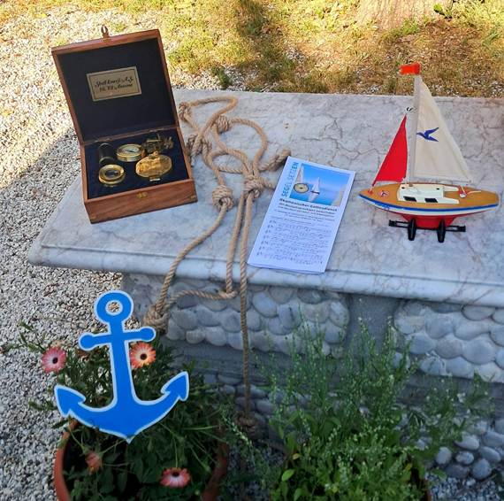 The image shows a decorative display featuring a model boat with a red sail, a blue anchor, and a plaque with a religious service title, all set on a pebbled surface with green foliage in the background.

AI-generated content may be incorrect.