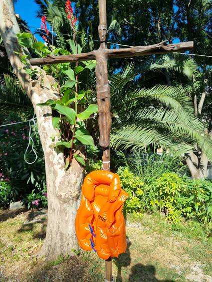 An orange life jacket hangs from a cross, surrounded by greenery and palm trees.

AI-generated content may be incorrect.