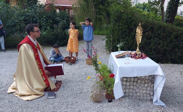 A person in a robe sitting on a gravel surface with a table with flowers and a gold object with a cross on it

Description automatically generated