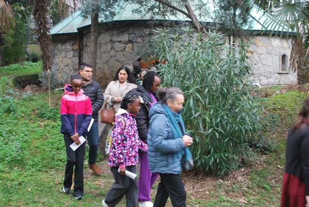 A group of people, including children in colorful clothing, is gathered around a rustic stone building in a garden setting.

AI-generated content may be incorrect.