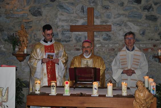 The image depicts a religious ceremony with three individuals dressed in ceremonial attire, standing around a table with a cross, a holy book, and various liturgical items, all set against a stone wall with decorations.

AI-generated content may be incorrect.