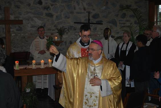 The image depicts a person dressed in a white alb and red sash, holding a lit candle, standing in front of a stone wall with a church setting and other people in religious attire.

AI-generated content may be incorrect.